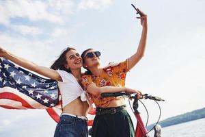 två patriotisk glad kvinnor med cykel och USA flagga i händer gör selfie foto