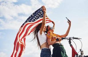 två patriotisk glad kvinnor med cykel och USA flagga i händer gör selfie foto