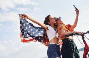 två patriotisk glad kvinnor med cykel och USA flagga i händer gör selfie foto