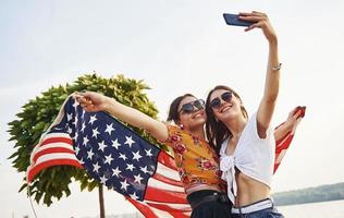 grön träd på bakgrund. två patriotisk glad kvinnor med USA flagga i händer framställning selfie utomhus i parkera foto