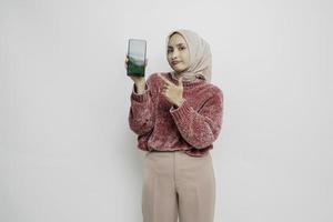 upphetsad asiatisk muslim kvinna bär rosa Tröja och hijab pekande på de kopia Plats bredvid henne medan innehav henne telefon, isolerat förbi vit bakgrund foto