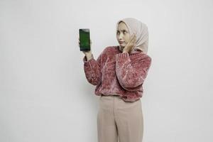 överraskad asiatisk muslim kvinna bär rosa Tröja och hijab som visar kopia Plats på henne smartphone, isolerat förbi vit bakgrund foto