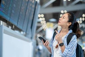 ung kvinna resande innehav cell telefon är stående ser på ett flygplats information styrelse i de flygplats. till kolla upp de flyg tid av henne egen. reser begrepp foto