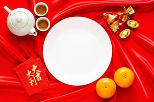 vit tallrik på röd satin trasa bakgrund med te uppsättning, tackor, röd väska ord betyder rikedom, apelsiner och röd kuvert paket eller ang bao ord betyder beskydd för kinesisk ny år begrepp. foto