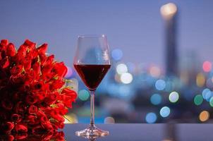 en glas av röd vin och röd ro bukett på tabell med färgrik stad bokeh lampor för årsdag eller valentine dag begrepp. foto