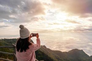 ung kvinna resenär som tar foto med smart telefon vid havet av dimma och soluppgång över berget