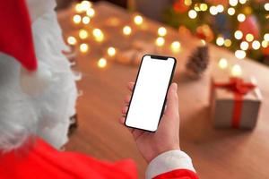 telefon attrapp i santa claus händer. jul lampor och gåvor i bakgrund. tom skärm för kopia eller produkt presentation foto