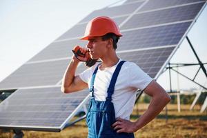 sladdlös skruvmejsel i hand. manlig arbetstagare i blå enhetlig utomhus med sol- batterier på solig dag foto