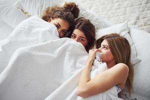 liggande på de säng under de filt tillsammans. Lycklig kvinna vänner har Bra tid på pyjamas fest i de sovrum foto