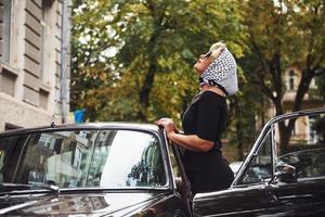 blond kvinna i solglasögon och i svart klänning nära gammal årgång klassisk bil foto
