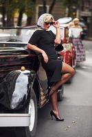 blond kvinna i solglasögon och i svart klänning lutande på gammal årgång klassisk bil foto