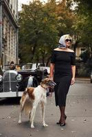 blond kvinna i solglasögon och i svart klänning nära gammal årgång klassisk bil med henne hund foto