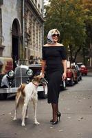 blond kvinna i solglasögon och i svart klänning nära gammal årgång klassisk bil med henne hund foto