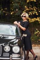 blond kvinna i solglasögon och i svart klänning nära gammal årgång klassisk bil foto