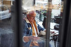 eleganta senior i modern kläder och i glasögon sitter i de Kafé och läser tidning foto