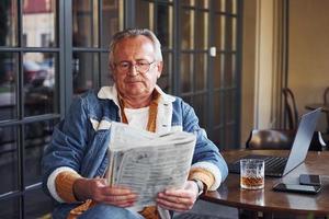 eleganta senior i modern kläder och i glasögon sitter i de Kafé och läser tidning foto