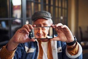 eleganta senior i modern kläder och i glasögon sitter i de Kafé och innehar telefon mot hans ansikte foto