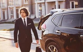 porträtt av stilig ung affärsman i svart kostym och slips utomhus nära modern bil i de stad foto