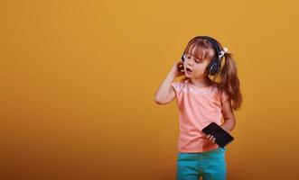 porträtt av söt liten flicka i hörlurar och med telefon i de studio mot gul bakgrund foto