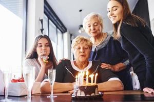 blåser de ljus. senior kvinna med familj och vänner fira en födelsedag inomhus foto