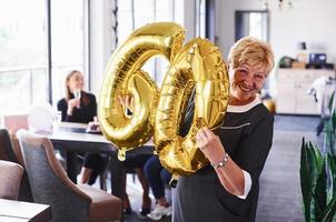 med ballonger av siffra 60 i händer. senior kvinna med familj och vänner fira en födelsedag inomhus foto
