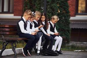 skola barn i enhetlig den där sitter utomhus på de bänk med smartphones foto