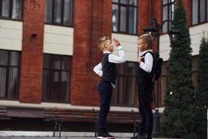 två liten Pojkar i skola enhetlig den där är utomhus tillsammans ge hög fem nära utbildning byggnad foto