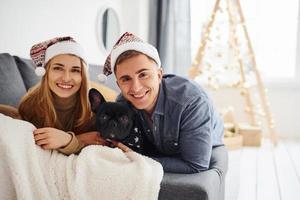 Lycklig ung modern par i jul hattar liggande ner på soffa på Hem med deras söt hund foto