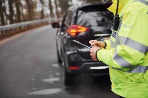 manlig polis officer i grön enhetlig stående med anteckningsblock nära bil foto