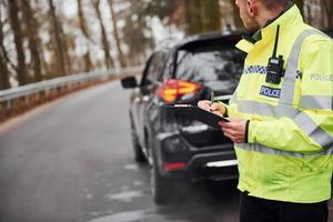 manlig polis officer i grön enhetlig stående med anteckningsblock nära bil foto