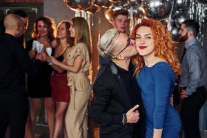 ung man kyssar rödhårig kvinna mot deras vänner i jul dekorerad rum och fira ny år foto
