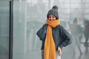 skön glad flicka i gul scarf och i värma kläder stående inomhus mot bakgrund med reflektioner foto
