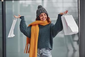 leende och känsla Lycklig. skön glad flicka i gul scarf och i värma kläder stående inomhus med handla påsar i händer foto