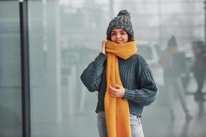 skön glad flicka i gul scarf och i värma kläder stående inomhus mot bakgrund med reflektioner foto