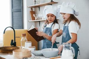 två liten flickor i blå kock enhetlig framställning mat på de kök och läsning mottagande bok foto