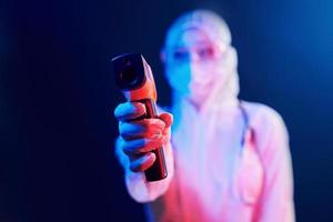 sjuksköterska i mask och vit enhetlig stående i neon upplyst rum och innehav infraröd termometer. sluta coronavirus spridning foto