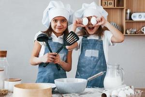 två liten flickor i blå kock enhetlig ha roligt medan framställning mat på de kök foto