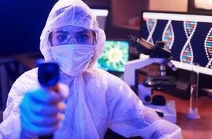 sjuksköterska i mask och vit enhetlig, innehav infraröd termometer och Sammanträde i neon upplyst laboratorium med dator och medicinsk Utrustning sökande för coronavirus vaccin foto