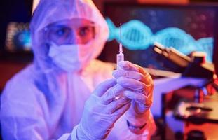sjuksköterska i mask och vit enhetlig, innehav spruta och Sammanträde i neon upplyst laboratorium med dator och medicinsk Utrustning sökande för coronavirus vaccin foto