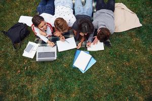 skrivning på papper. med bärbar dator. grupp av ung studenter i tillfällig kläder på grön gräs på dagtid foto