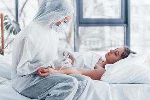 kvinna läkare i defensiv labb täcka och skyddande glasögon med spruta i hand injicering medicin till ung flicka sjuk av virus foto