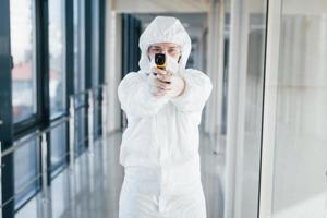 kvinna läkare forskare i labb täcka, defensiv glasögon och mask stående inomhus med infraterad termometer foto