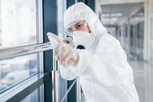 kvinna läkare forskare i labb täcka, defensiv glasögon och mask stående inomhus med antibakteriell spray foto