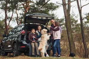 Lycklig familj ha roligt med deras hund nära modern bil utomhus i skog foto