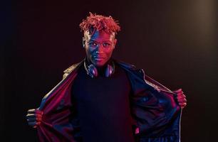 Framställ med trådlös hörlurar på nacke. trogen neon belysning. ung afrikansk amerikan man i de studio foto
