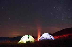 två upplyst tält med lägereld under stjärnor på bergen på natt foto