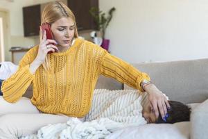 mor mätning temperatur av henne sjuk unge. sjuk barn med hög feber om i säng och mor innehav termometer. mor med cell telefon kallelse till läkare foto