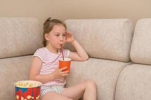 söt flicka med en hårpiska sitter på Hem på de soffa tittar på en film med entusiasm och dricka cola, Nästa till en glas av popcorn foto