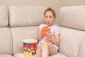 söt liten flicka sitter på Hem på de soffa med en enorm glas av popcorn och drycker en cocktail genom en sugrör, Nästa till de avlägsen kontrollera från de TV foto