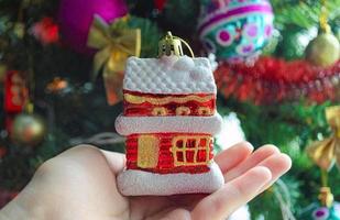 hand innehav jul träd leksak i de form av liten hus. verklig egendom mäklare jul. foto